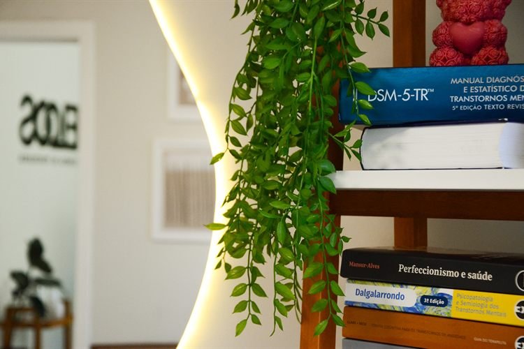 A estante do consultório, com livros de psicologia, uma planta pendente e a luz do espelho acesa atrás.